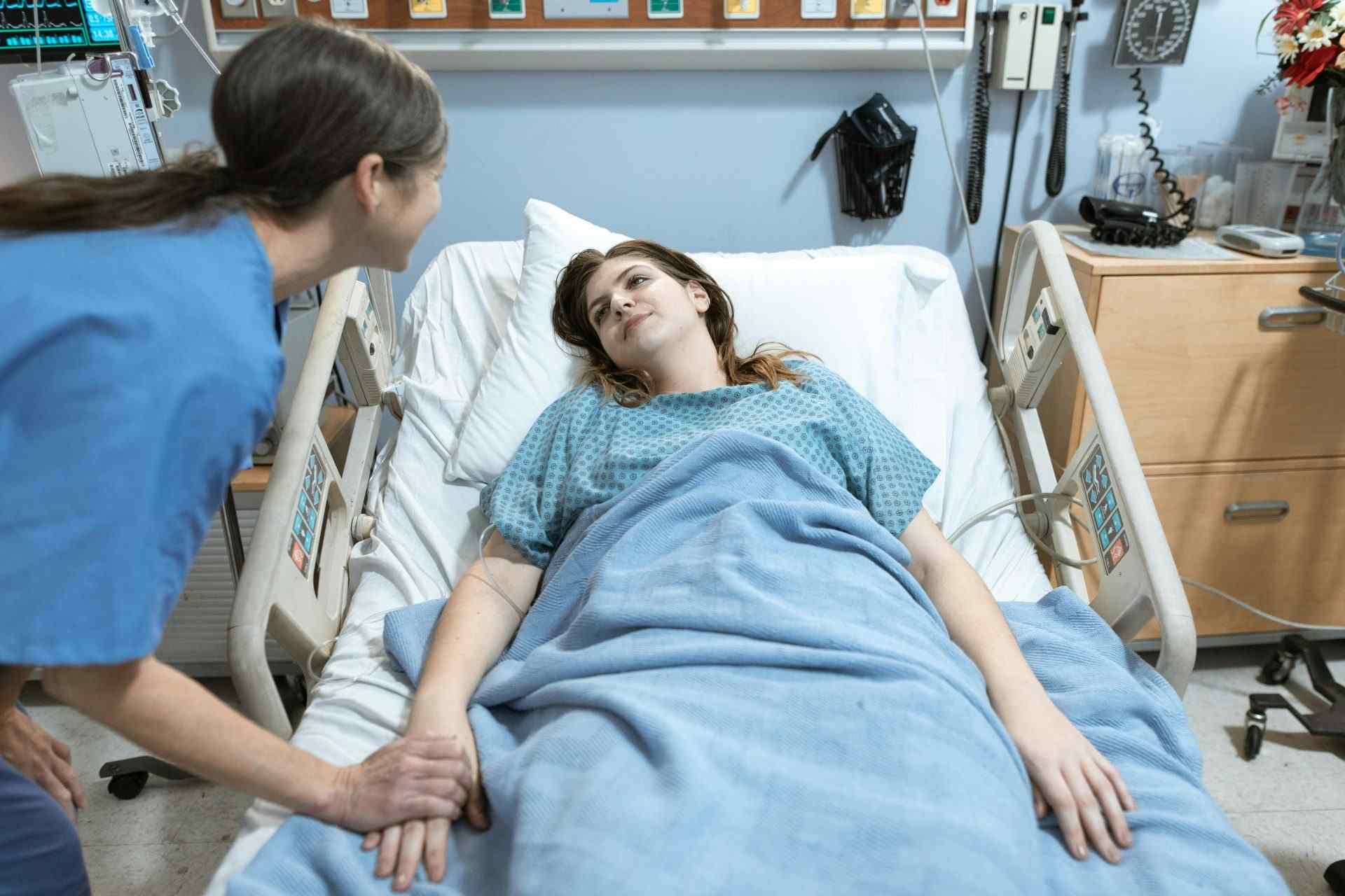 Krankenschwester in blauen Kasacks spricht mit einer jungen Patientin, die in einem Krankenhausbett liegt